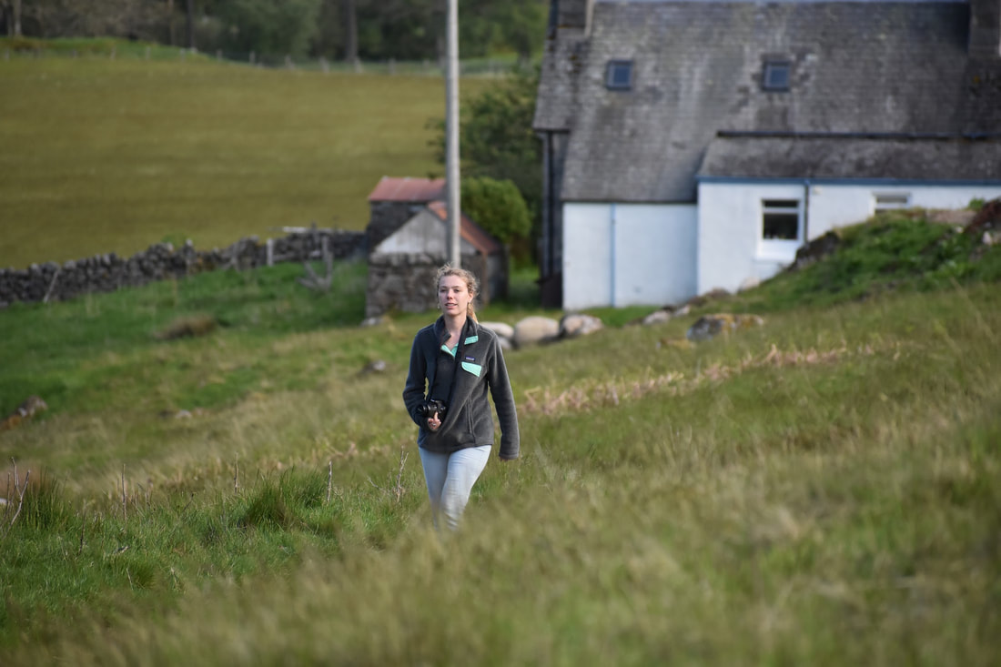 Erin, a woman with curly blond hair in a ponytail, walks through a field of grass in front of a farmhouse.