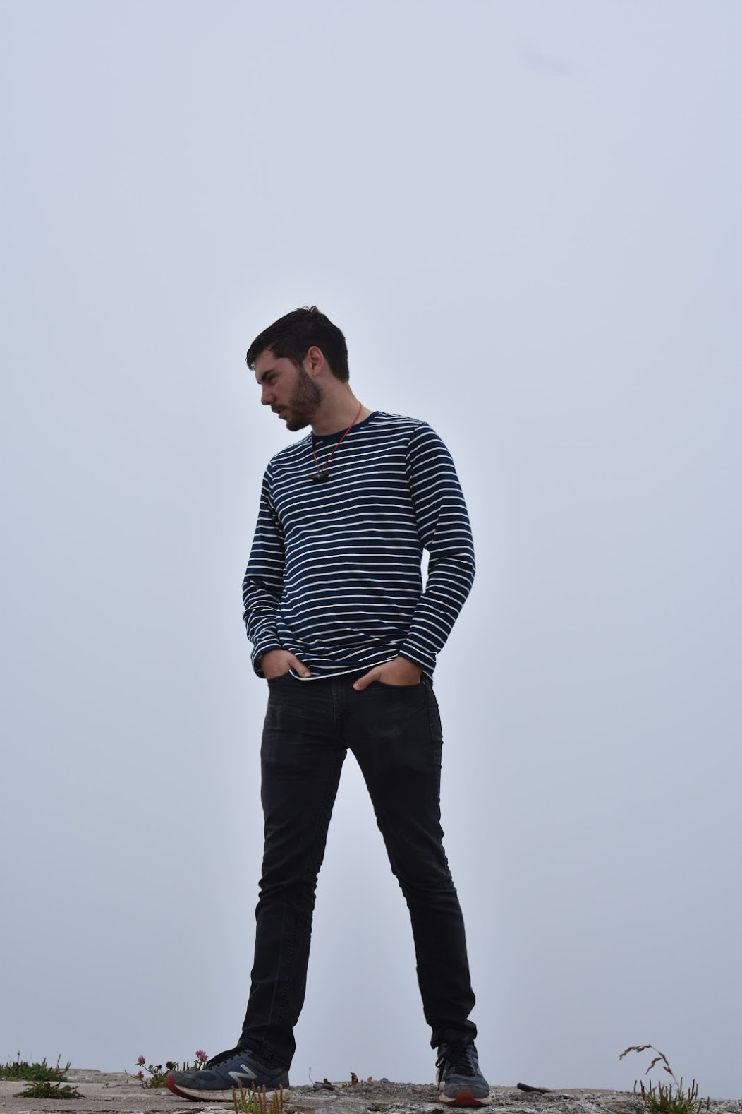 My brother Nathan, with short dark hair and a beard, wearing a black-and-white long sleeved striped shirt. He strikes a hands-in-pockets pose against a gray foggy sky.