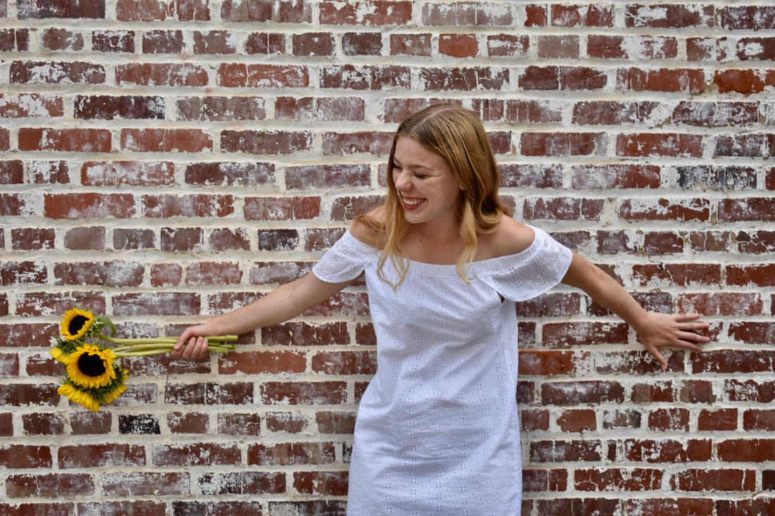 Erin, with blonde hair and wearing a white off-the-shoulder dress, stands in front of a faded brick wall. She smiles and looks away, holding sunflowers in her right hand.