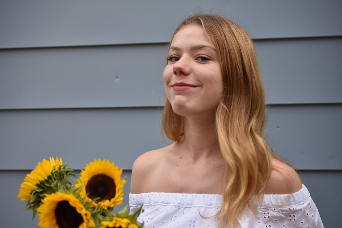 Erin, a white woman with blonde hair and a white dress, stands confidently in front of a teal siding, sunflowers in hand.