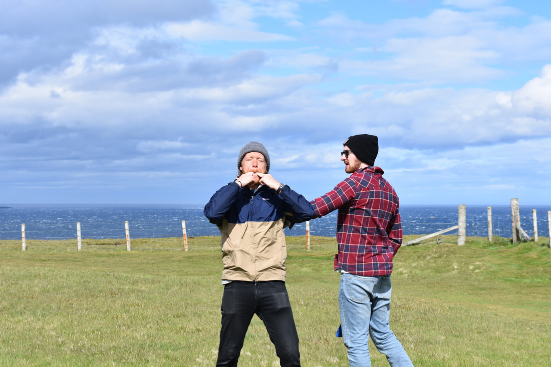 Henry stands in a blue-and-khaki jacket, preparing to let out a fierce two-fingered whistle. Eric, in a black cap and red plaid shirt, moves to stop him. They both stand in some grass with a fence and the ocean behind them.