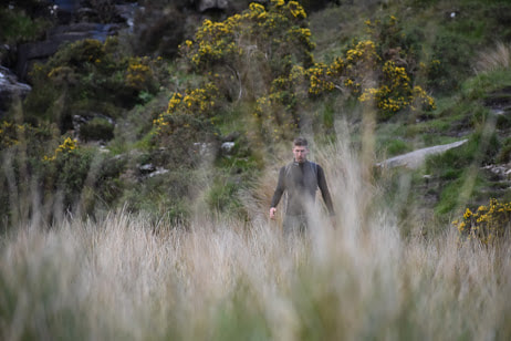 My friend Lucas, wearing a black shirt, walks through a field of grass. A mountainside rises behind him, covered in gorse, shrubs, and rock.
