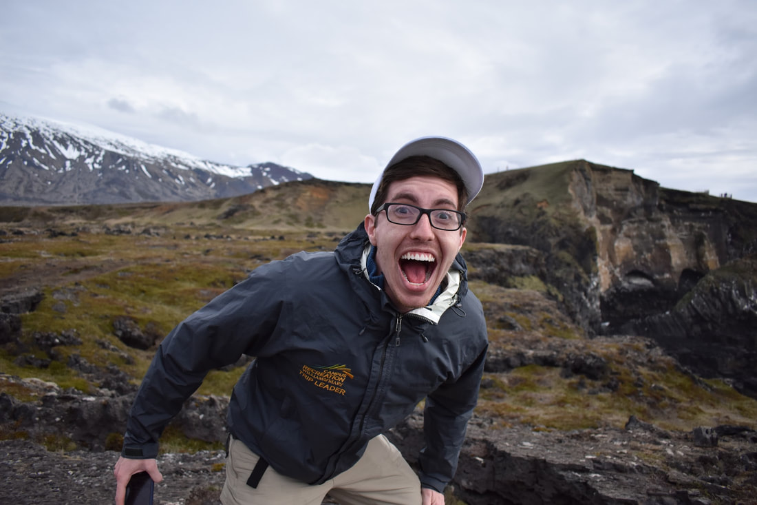 Ian expresses wonder at the Icelandic coastline, mouth agape and eyes wide behind his glasses. He wears a windbreaker and a gray rainslicker.
